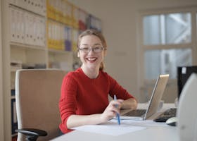 pexels-photo-3783725-3783725 Happy woman in red sweater working at office desk with laptop and pen, smiling at camera.